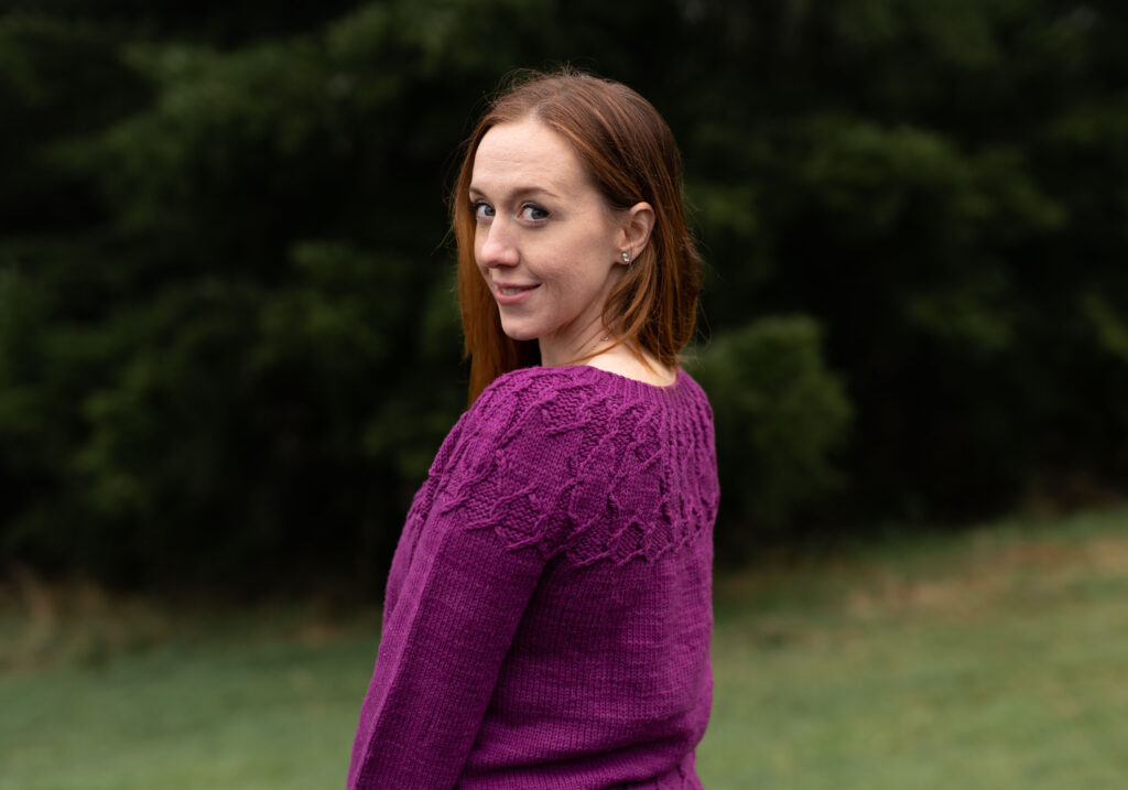 Side View of smiling woman wearing the Alcyone sweater in front of dark green evergreen trees copyright Avi Dascaloff 2020