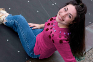 Seated woman smiling at camera over shoulder wearing fitted Circle Dance Sweater digitally altered to appear knit in a pink Space Cadet colorway