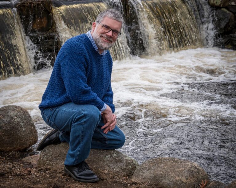 Man wearing Herring Run Sweater kneels by a stream and waterfall full of herring