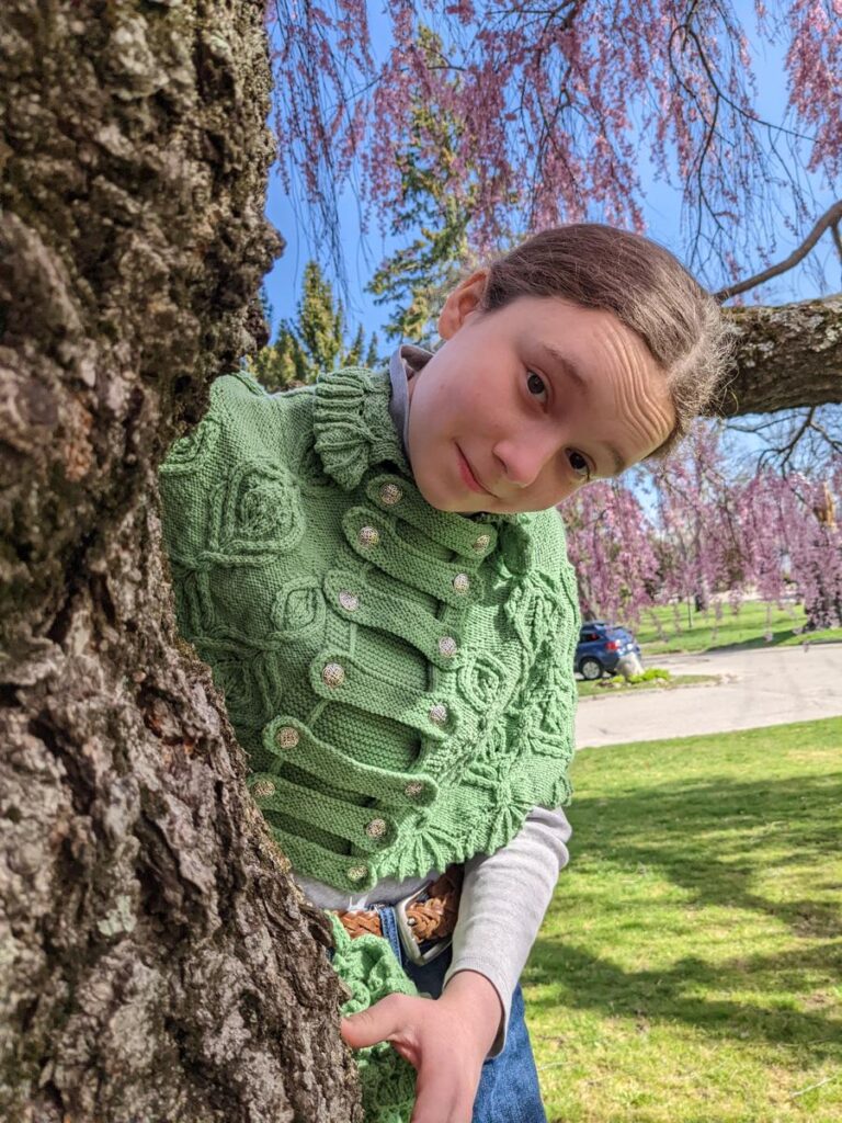 Girl wearing the Silvia of ELF cape peeks around the trunk of a cherry tree