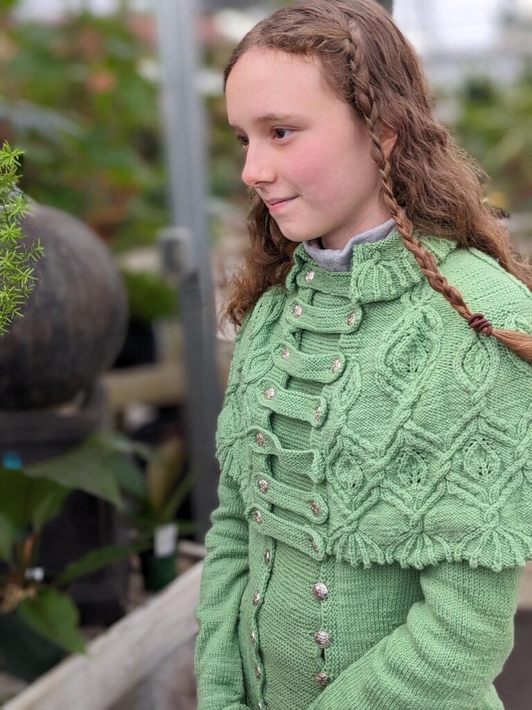 Young woman models the Silvia Got Promoted Sweater with the Silvia of ELF cape buttoned onto it.