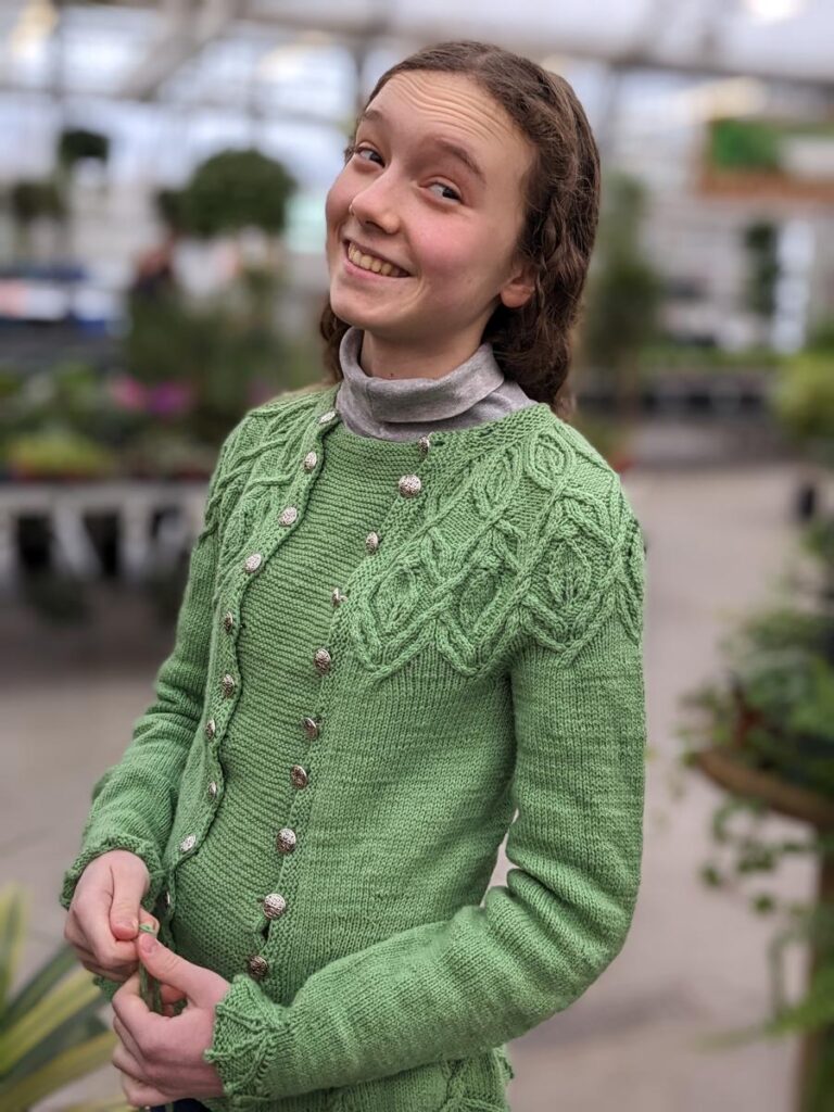 Young woman models the Silvia Got Promoted Sweater in light green, in a green house.