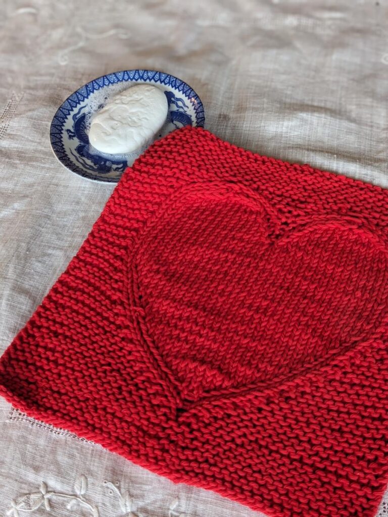 Merry Heart facecloth worked in red yarn, displayed on a white cloth, with dish of soap